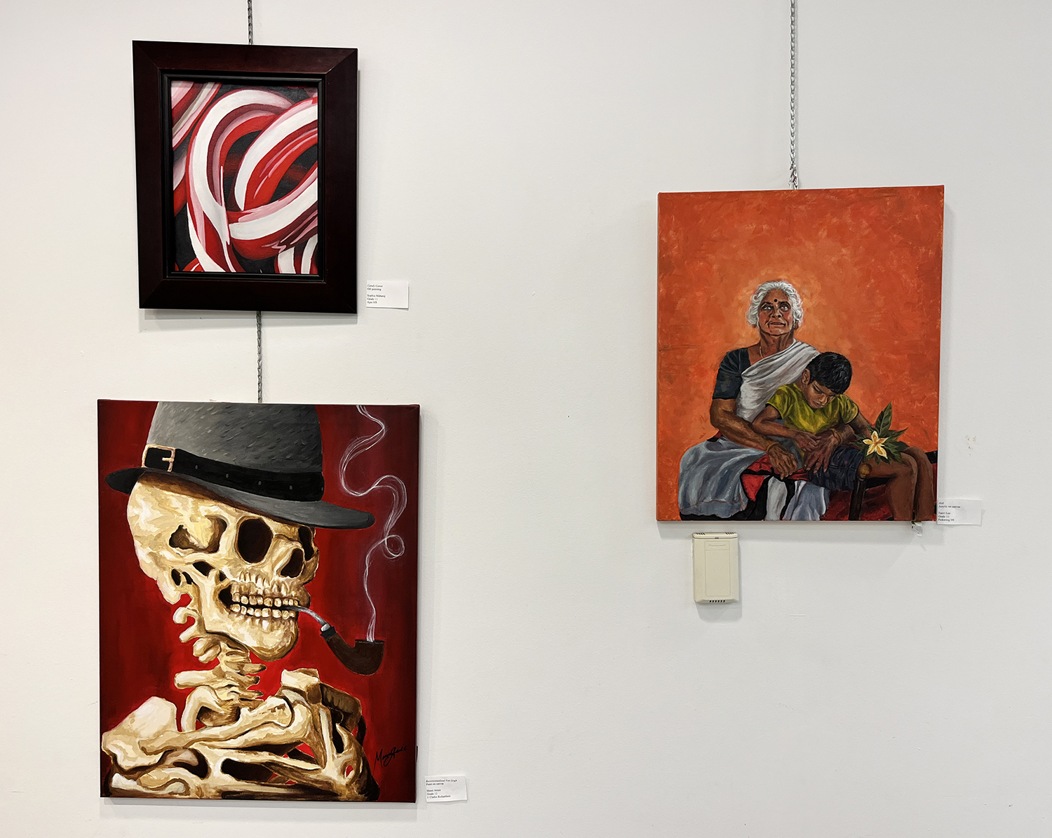 Three paintings from the Heartbeat Art Show hang on a wall in the Rotary Room at Main Branch. One painting is red and white swirls, one painting is of a skeleton wearing a hat, and the other is of a person hugging a child.